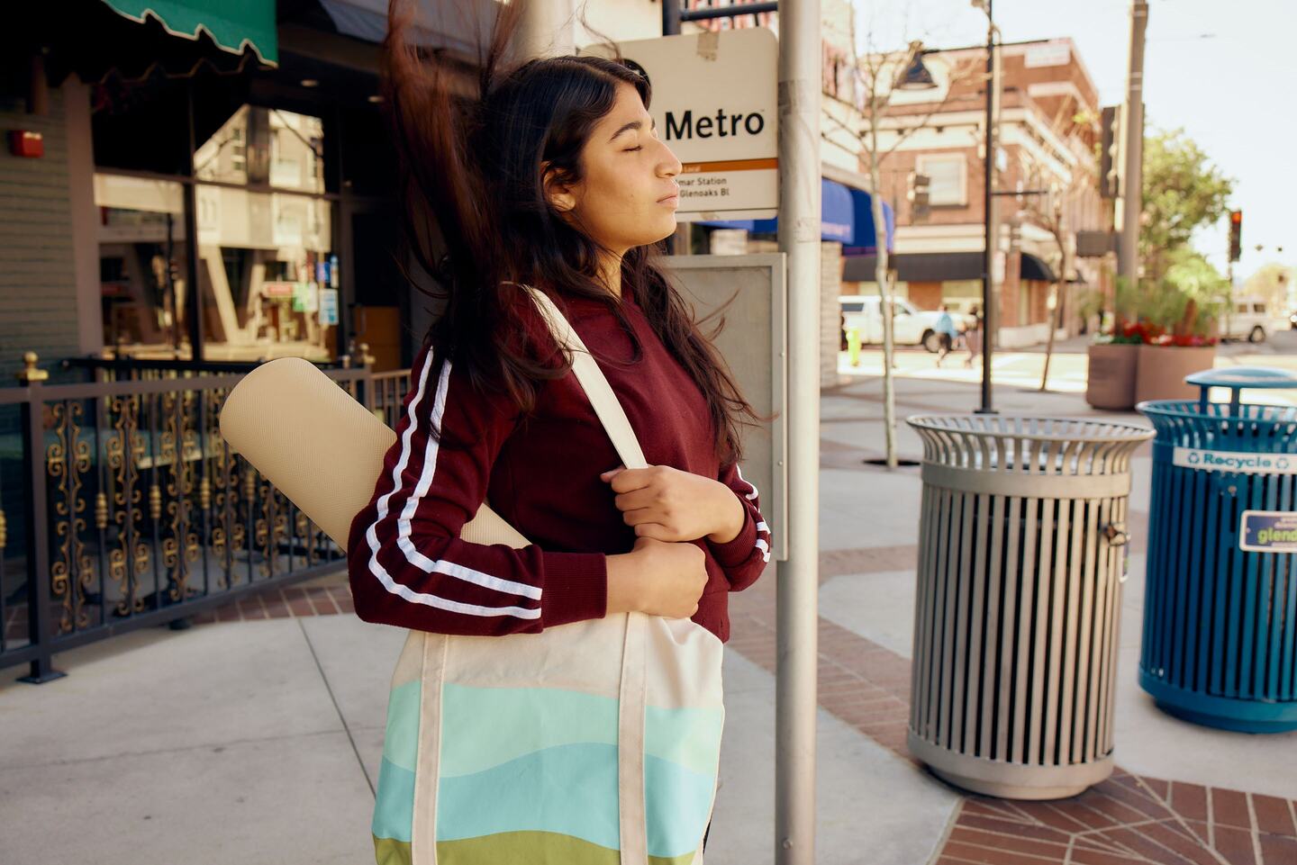 Woman with a rolled yoga mat over her shoulder, eyes closed, standing on a city sidewalk